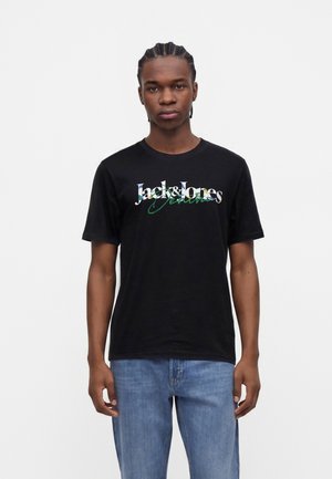 Homme aux cheveux tressés portant un t-shirt noir Jack & Jones et un jean bleu, debout devant un fond blanc.