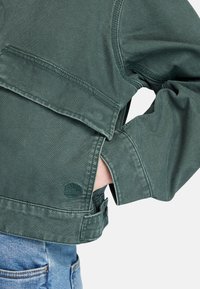 Groene spijkerjack met een oversized pasvorm, voorzien van een achterzak en een logo-embleem. Textuur lijkt zacht met zichtbare stiksels.