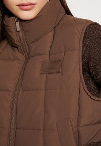 Gilet imbottito marrone con colletto alto, dotato di chiusura con zip e un piccolo patch in pelle, indossato sopra un maglione marrone a trama.