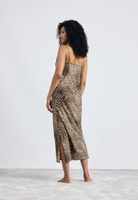 Leopardmønster satin glidkjole med tynne stropper, splitt i ryggen og glatt tekstur, som faller under kneet. Modell som står barføtt på en lys gulv.