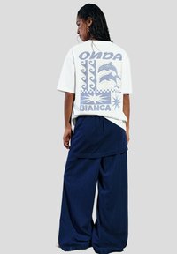 T-shirt blanc oversize avec un design graphique bleu présentant des dauphins, des vagues et des motifs géométriques, associé à un pantalon bleu marine décontracté.