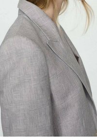 Blazer a cuadros grises hecho de un tejido texturizado; cuenta con un cuello de muesca, hombros estructurados y costuras visibles a lo largo de los bordes.