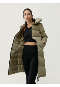 Olivgrön lång puffjacka med huva, med dragkedjestängning och elastiska detaljer. Bärs över en svart träningsskjorta och leggings.