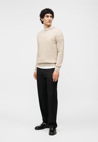 Mann steht, trägt einen beigen Langarmpullover, ein weißes Unterhemd, schwarze Hose und schwarze Schuhe, blickt nach links vor einfachem Hintergrund.