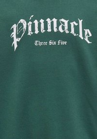 Zielona koszulka z białym tekstem w stylu gotyckim, który głosi "Pinnacle" i "Three Six Five", o gładkiej, miękkiej fakturze.