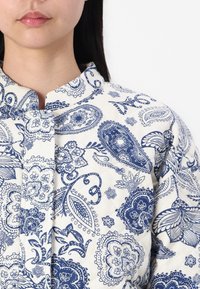 Biała pikowana koszula z tkaniny z niebieskim wzorem paisley, wyposażona w stójkę i zapięcie na guziki, z misternymi kwiatowymi detalami.
