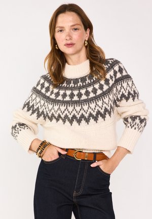 Maglione lavorato a maglia in crema con un collo patito grigio, maniche lunghe, polsini a coste e un collo aderente, abbinato a jeans scuri e una cintura marrone.