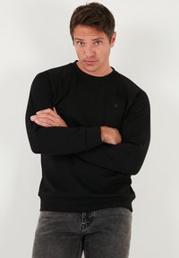 Fekete pulóver pamut keverékből, kerek nyakkivágással, hosszú ujjakkal, bordázott mandzsettákkal és szegéllyel, kis logó részlettel a mellkason.