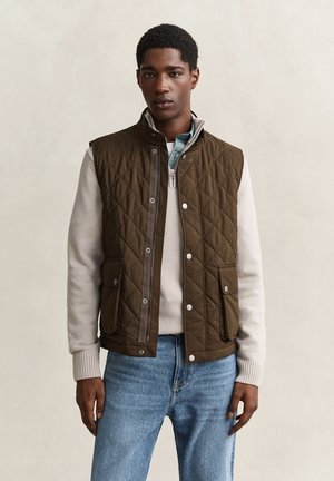 Junger Mann trägt eine braune gesteppte Weste über einem hellbeigen Pullover und blauen Jeans, steht vor einem einfarbigen neutralen Hintergrund.