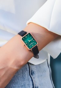 Rektangulär klocka med en grön texturerad urtavla, guldkantade detaljer och arabiska siffror, fäst vid ett svart läderarmband.