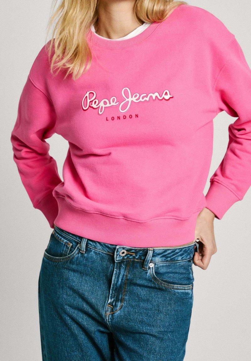 Blondýna oblečená v jasně růžovém svetru Pepe Jeans London a modrých džínách, s rukama zastrčenýma v pase džínů.