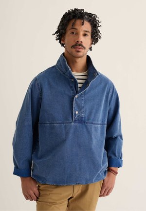 Hombre con cabello rizado y bigote que lleva un pulóver de mezclilla azul con botones a presión y mangas remangadas sobre una camisa a rayas, posando frente a un fondo beige.