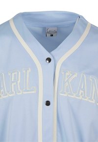 Maglia sportiva azzurro chiaro con accenti bianchi, caratterizzata dal testo "KARL" e una chiusura con bottoni. Realizzata in tessuto mesh traspirante.