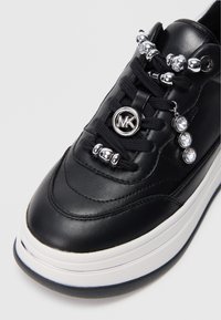 Mustast nahast tennisekad, millel on hõbedast detailid, sealhulgas dekoratiivsed pärlid ja kolm sädelevat kivi. Omab valget vaheosa ja tekstureeritud keelt.