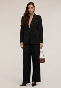 Completo nero su misura con giacca a un bottone, pantaloni larghi e tacchi a punta. Caratterizzato da dettagli dorati e una borsa bordeaux.