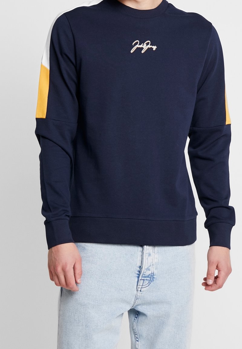 Uomo che indossa un felpa navy con pannelli bianchi e gialli sulle maniche e jeans azzurri chiari, in piedi contro uno sfondo bianco.