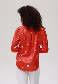Blusa di raso rosso con maniche lunghe, motivo floreale in bianco, giallo e nero, orlo curvato, dettaglio piega sul retro, texture liscia.