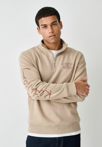 Beige sweatshirt med halv lynlås lavet af blødt stof. Har et dekorativt logo på brystet og tekst langs venstre ærme. Ribstrikkede manchetter og kant.