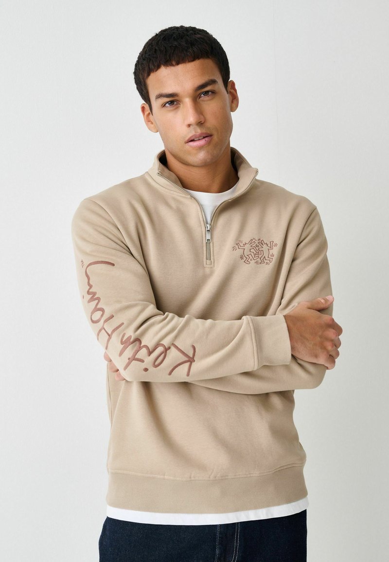 Beige sweatshirt med halv lynlås lavet af blødt stof. Har et dekorativt logo på brystet og tekst langs venstre ærme. Ribstrikkede manchetter og kant.