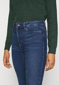 Höga blå jeans med subtil blekning, metallknappstängning och femfickorsdesign, ihop med en mörkgrön stickad tröja.