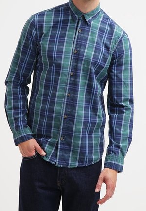 Homme portant une chemise à carreaux bleue et verte à boutons avec manches longues et un jean bleu foncé, une main dans la poche.