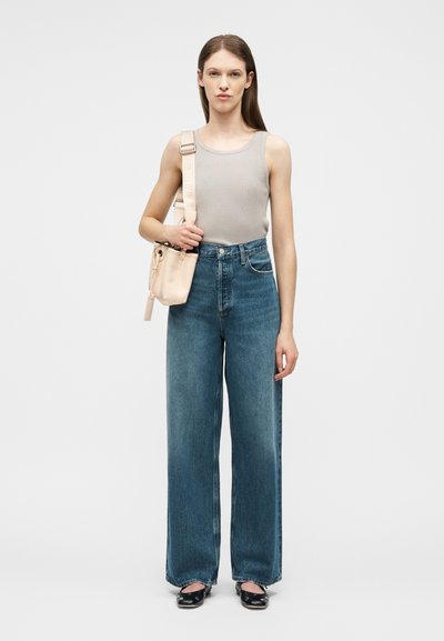 Γυναίκα που φορά γκρι αμάνικη μπλούζα, τζιν παντελόνι με ψηλή μέση και φαρδιά μπατζάκια σε μπλε χρώμα, μαύρα παπούτσια και κρατά μια μπεζ τσάντα ώμου σε λευκό φόντο.