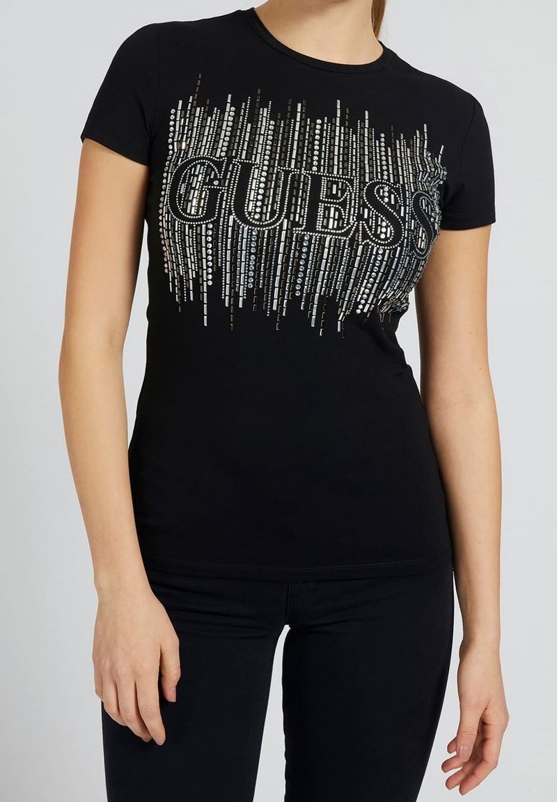 T-shirt noir ajusté avec des lignes verticales cloutées argentées et un grand logo "GUESS" sur la poitrine, porté par une personne debout.