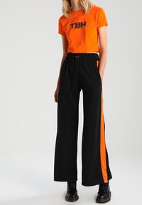 Mujer vestida con una camiseta corta naranja que dice "TBH", pantalones anchos negros con rayas laterales naranjas y botas negras con cordones.