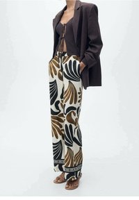 Blazer marron sur un crop top en tricot noir, associé à un pantalon large avec de grands motifs de feuilles noires et marron sur un fond crème.