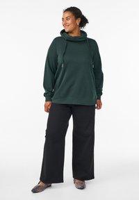 Pullover hoodie verde scuro con collo a cappuccio, maniche lunghe e cordini regolabili, abbinato a pantaloni neri a gamba larga e ballerine con stampa leopardata.