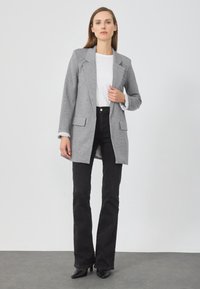 Blazer grigio con design su misura, due tasche frontali e spalle strutturate, indossato sopra una maglietta bianca e abbinato a jeans neri a zampa.