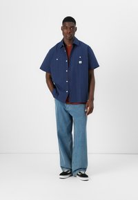 Kortermet marineblå striped skjorte med hvite knapper og brystlomme, kombinert med lyseblå vide jeans og svarte sneakers.