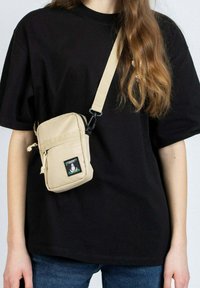 Beige olkalaukku, joka on valmistettu sileästä kankaasta. Sisältää etupussin ja värikkään logopatchin. Säädettävä olkahihna mukana.