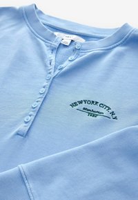 Chemise henley bleu clair à manches longues avec boutons et texte brodé en vert indiquant New York City, N.Y. Manhattan 1985 sur la poitrine.