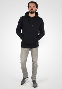 Sudadera negra con cordones y bolsillo frontal, combinada con jeans gris claro y zapatillas deportivas grises. Diseño simple, hecha de material suave.