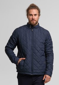 Gequilte donkerblauwe jas met een hoge kraag, rits aan de voorkant en drukknopen sluiting. Voorzien van zijzakken en een gestructureerde stof. Slanke pasvorm.