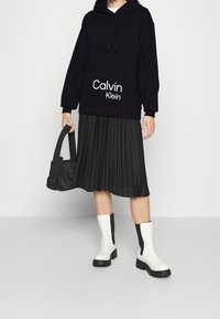 Osoba v černé mikině Calvin Klein, černé plisé sukni, bělostných botách po lýtka, držící černou kabelku na jednobarevném pozadí.