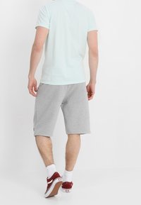 Mężczyzna stoi tyłem do aparatu, ubrany w jasnoniebieską koszulkę, szare szorty do kolan, białe skarpetki i czerwone trampki.