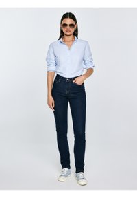 Sieviete, tērpusies gaiši zilā pogājamā kreklā, tumši zilos pieguļošos džinsos, gaiši zilās sporta kurpēs un saulesbrillēs, stāv pret vienkāršu fonu.