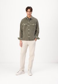 Olijfgroene corduroy jas met twee borstzakken, knoopsluiting en ingeplooide kraag, gecombineerd met beige broek en witte sneakers.