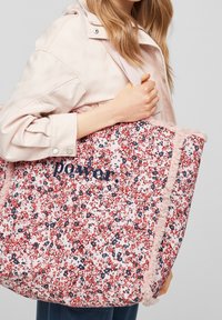 Kobieta trzymająca dużą torbę z kwiatowym wzorem, z różową frędzelką i wyszytym napisem "moc", ubrana w jasnoróżową kurtkę i ciemne spodnie.