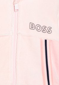 Veste en polaire rose clair avec une fermeture éclair à l'avant, des rayures noires et roses sur les côtés, et le logo "BOSS" en fil noir sur la poitrine.