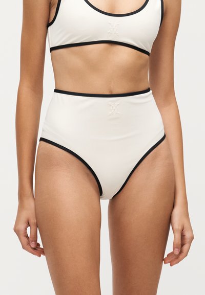 Femme portant un bas de bikini blanc taille haute et un haut assorti avec une bordure noire sur fond uni.