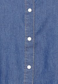 Tessuto denim di colore blu, con bottoni bianchi e cuciture a contrasto lungo la pattina. Texture liscia, design casual.