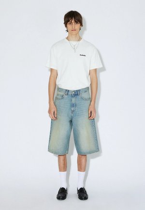 Jonge man draagt wit T-shirt met korte mouwen, lichtblauwe spijkershorts tot op de knie, witte sokken en zwarte loafers, staand tegen een witte achtergrond.