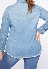 Ljust blå denimskjorta med spetsig krage, långa ärmar och fransad fåll; har sömnadsdetaljer på axlarna och ryggen.