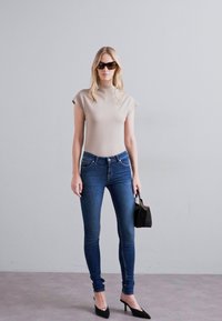 Top beige aderente con maniche a cappuccio, jeans skinny blu, scarpe con tacco nere e una piccola borsa nera, su uno sfondo neutro.