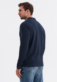 Man med kort brunt hår som bär en marinblå långärmad skjorta och blå jeans, som ser åt vänster mot en vit bakgrund.