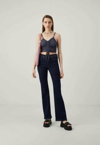 Nuori nainen pukeutuneena tummansiniiseen rypytettyyn toppiin, tummiin leveälahkeisiin farkkuihin, mustiin korokepantterisaappaisiin, värikkääseen helmiketjuun ja kantaa vaaleanpunaista kudottua olkalaukkua.