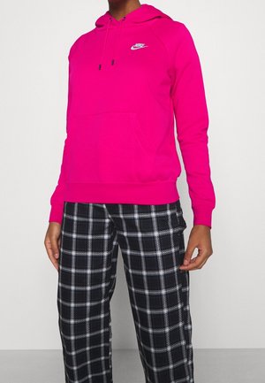 Persoon met felroze Nike-hoodie en zwart-witte geruite broek, staand tegen een effen lichte achtergrond.